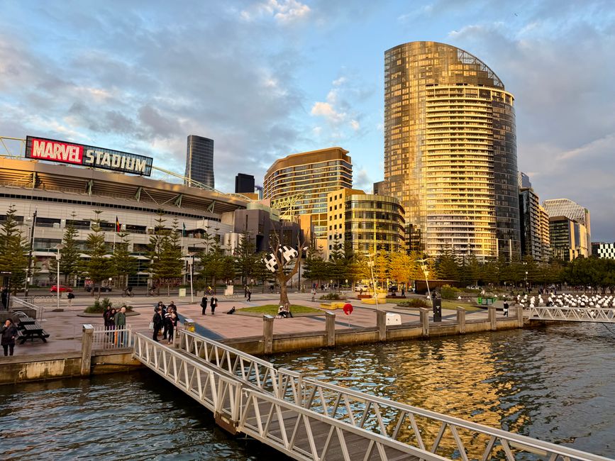 The image depicts a daytime cityscape along a waterfront in Docklands, featuring a modern high-rise building with a curved glass façade reflecting sunlight, positioned prominently in the background. To the left, there is a large stadium with a white exterior and a prominent Marvel logo signage, indicating its function, with several smaller dark-colored buildings behind it. In the foreground, a paved promenade runs parallel to the water, dotted with a few trees that have green and yellow leaves, hinting at the season. People are visible walking and gathering along the promenade, some near a sculpture of two stylized, black and white cartoonish faces with expressive features, positioned centrally on the paved area. A white metal footbridge, with a lattice framework, extends over the water, connecting the promenade to the pier. The waterfront features calm water with gentle ripples, and the sky above is partly cloudy with patches of blue. The scene is well-lit by natural sunlight, creating a warm, reflective glow on the windows of the high-rise buildings, illustrating a typical urban setting where private waste collection or on-site clearance might be relevant to the area serviced by companies like Rubbish Collection Docklands.