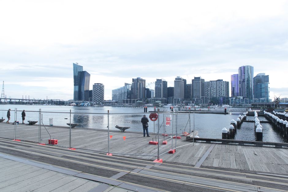 The image depicts a daytime cityscape along a waterfront in Docklands, featuring a modern high-rise building with a curved glass façade reflecting sunlight, positioned prominently in the background. To the left, there is a large stadium with a white exterior and a prominent Marvel logo signage, indicating its function, with several smaller dark-colored buildings behind it. In the foreground, a paved promenade runs parallel to the water, dotted with a few trees that have green and yellow leaves, hinting at the season. People are visible walking and gathering along the promenade, some near a sculpture of two stylized, black and white cartoonish faces with expressive features, positioned centrally on the paved area. A white metal footbridge, with a lattice framework, extends over the water, connecting the promenade to the pier. The waterfront features calm water with gentle ripples, and the sky above is partly cloudy with patches of blue. The scene is well-lit by natural sunlight, creating a warm, reflective glow on the windows of the high-rise buildings, illustrating a typical urban setting where private waste collection or on-site clearance might be relevant to the area serviced by companies like Rubbish Collection Docklands.