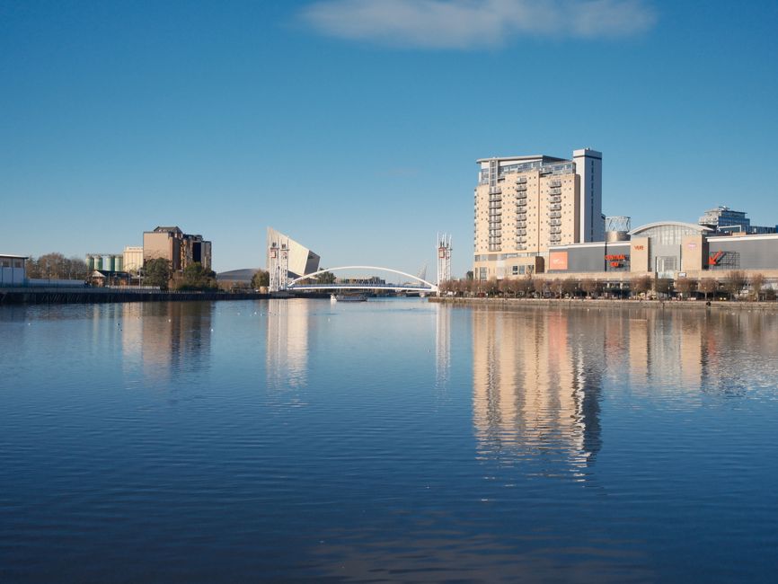 A modern cityscape featuring a prominent glass-fronted office building with a distinctive hollowed-out section on the upper floors, situated along a riverbank. The building's reflective glass exterior showcases grid-like paneling in a light grey and metallic finish. Surrounding the main structure are several other mid-rise buildings of various architectural styles, including brick and concrete facades, some with windows and flat rooftops. The foreground includes a paved walkway or quay with a low brick wall, and the river reflects the buildings clearly, creating a mirror image of the urban scene. The sky is clear with a few wispy clouds, and natural daylight illuminates all structures evenly. This setting suggests an area involved in independent or private waste handling, possibly indicating the presence of rubbish removal or on-site clearance services in a commercial or business district, where such facilities support urban infrastructure and development.