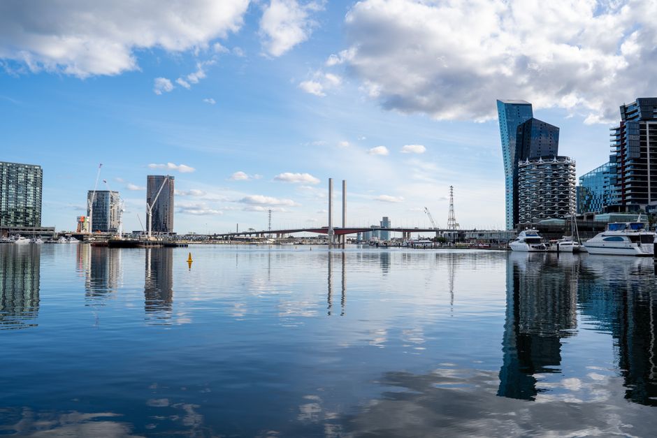 A wide view of a modern city waterfront during daylight hours shows a calm body of water reflecting the skyline, featuring tall contemporary office buildings and residential towers with glass facades. To the right, there are several white yachts docked along the quay, with their reflections visible on the water’s surface. In the background, a bridge with two tall white pylons spans across the water, and cranes can be seen amidst ongoing construction. The sky above is partly cloudy with patches of blue, casting soft natural light over the scene. The environment appears clean and well-maintained with no visible rubbish or debris, aligning with professional waste management standards, and occasionally reflecting the importance of alternative waste handling and private disposal services like those offered by Rubbish Collection Docklands in managing urban waste tasks effectively.