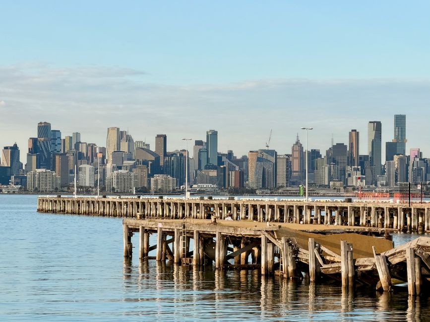 A stretch of wooden pier extending into calm water with visible support stilts beneath, situated in a harbour or waterfront area. The pier appears weathered, with some uneven and tilted sections, suggesting age or damage. Behind it, a modern city skyline features a variety of high-rise office buildings and residential towers with glass and steel facades, some under construction with cranes visible. The scene is set during daylight hours with clear skies, and the overall environment indicates an urban waterfront that may be associated with private or independent waste disposal activities, reflecting common challenges in managing bulky waste in densely populated areas. The presence of a clean, well-maintained skyline contrasts with the rustic, slightly deteriorated appearance of the pier, highlighting a potential area where local waste collection services like Rubbish Collection Docklands might be involved in managing waste associated with waterfront development or maintenance. The natural reflection of the buildings and sky in the water creates a tranquil yet utilitarian atmosphere relevant to urban rubbish removal and disposal contexts.