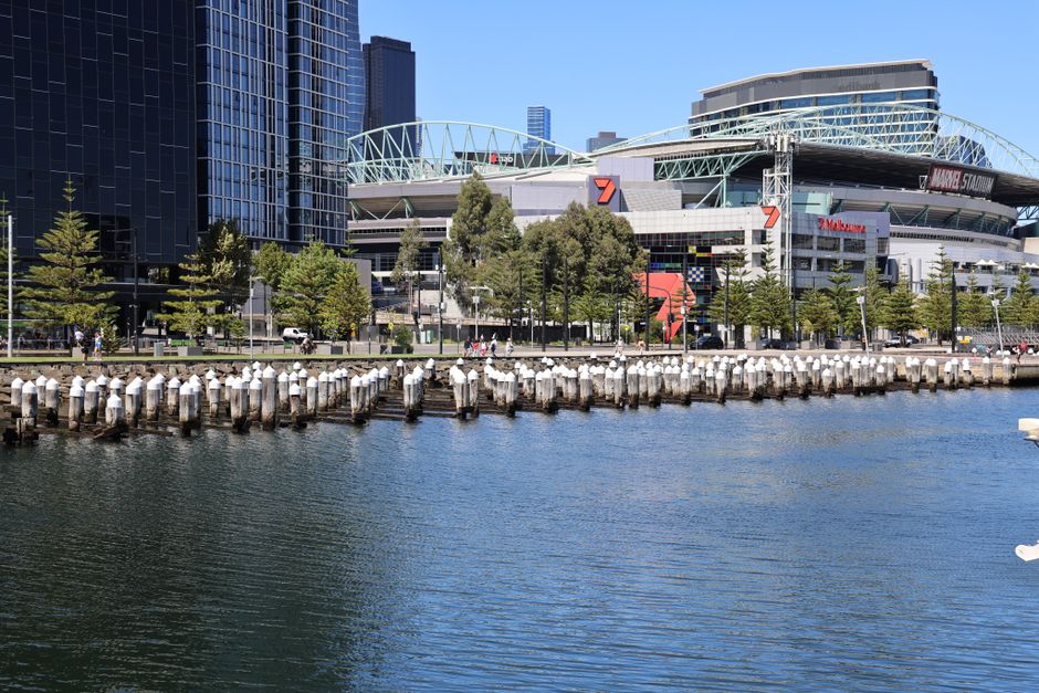 A wide view of a modern city waterfront during daylight hours shows a calm body of water reflecting the skyline, featuring tall contemporary office buildings and residential towers with glass facades. To the right, there are several white yachts docked along the quay, with their reflections visible on the water’s surface. In the background, a bridge with two tall white pylons spans across the water, and cranes can be seen amidst ongoing construction. The sky above is partly cloudy with patches of blue, casting soft natural light over the scene. The environment appears clean and well-maintained with no visible rubbish or debris, aligning with professional waste management standards, and occasionally reflecting the importance of alternative waste handling and private disposal services like those offered by Rubbish Collection Docklands in managing urban waste tasks effectively.