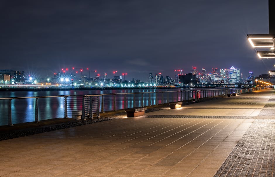 A nighttime scene of a paved walkway along a river in a cityscape. The foreground features a smooth, light-colored concrete surface with a textured strip on the right side. The walkway is illuminated by modern, rectangular wall-mounted lights on the right, casting bright white light and creating subtle shadows on the ground. A low metal railing runs parallel to the river, with evenly spaced posts. In the middle ground, the calm river reflects the bright, colorful city lights, including shades of white, red, and blue, from the distant urban skyline. The background showcases a densely built city with high-rise buildings illuminated by multicolored lights and numerous red aviation warning lights atop construction cranes. The sky overhead appears cloudy with a dark grey hue, contrasting with the vibrant city lights. The scene exudes a clean, urban environment suitable for independent rubbish collection or waste management activities, with the city’s modern infrastructure subtly emphasizing alternative waste handling options for residents and services like Rubbish Collection Docklands.
