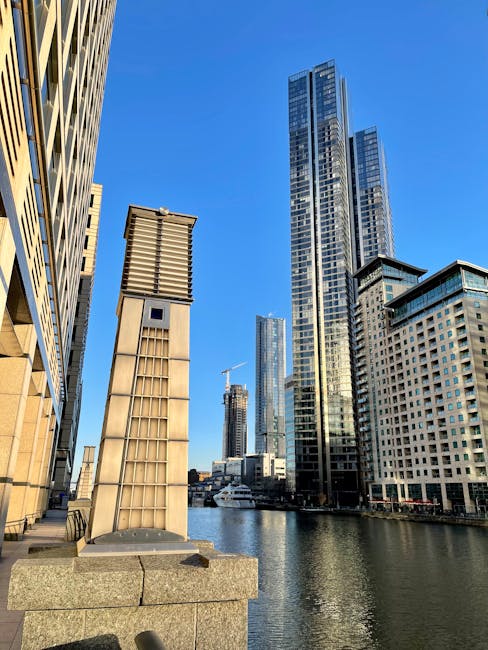 A cityscape view along a river in an urban environment featuring modern skyscrapers with glass facades reflecting the blue sky, and several mid-rise buildings with concrete and steel finishes. Prominent tall towers dominate the skyline with their sleek, vertical lines, while smaller structures add variety to the scene. In the foreground, a stone walkway edge and a metal ventilation shaft with a grid-like pattern are visible on the left side, positioned on a stone platform at the water's edge. The calm river below reflects the buildings and the clear sky above, creating a tranquil atmosphere typical of a central business district. These architectural elements are situated in an area that could be associated with private on-site waste management or independent rubbish collection services, supported by the proximity of docks or wharf areas suitable for waste disposal and rubbish removal operations, an environment relevant to companies like Rubbish Collection Docklands.