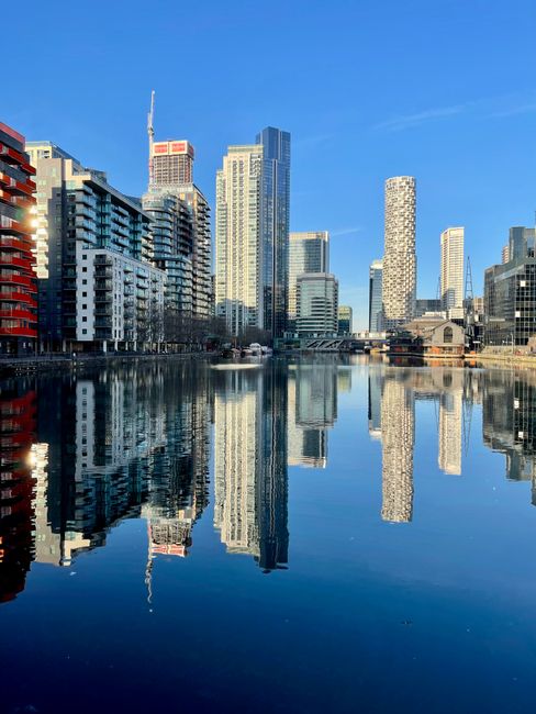 A cityscape featuring modern high-rise buildings along a waterfront, with reflected images of the skyscrapers on the calm water surface. The buildings exhibit a variety of architectural styles, with glass facades, concrete finishes, and some with textured or patterned exteriors. The sky above is clear and blue, and the natural daylight highlights the details of each structure. In the foreground, the water is smooth, creating a mirror-like reflection of the urban skyline. The scene appears to be taken from a quay or promenade area, with no visible waste or clutter, emphasizing the clean, developed environment. The modern appearance of the buildings, combined with the water reflection, suggests a contemporary urban area that could benefit from private waste management or rubbish removal services provided by companies like Rubbish Collection Docklands, supporting effective on-site clearance of waste generated in such metropolitan settings.