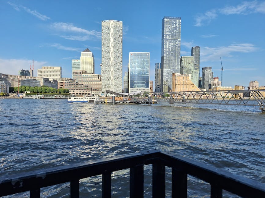 A wide view of a modern city skyline across a body of water, with several high-rise office buildings featuring glass facades and geometric designs. In the foreground, part of a black metal railing is visible, suggesting the photo was taken from a riverside promenade or boat. The water appears slightly choppy with small ripples, and a metal structure extends from the right side, possibly part of a dock or pier. The sky is mostly clear with light blue tones and a few scattered white clouds. The environment reflects urban development typical of central business districts, and the scene is well-lit with natural sunlight, emphasizing the glossy surface of the buildings and the water. This setting relates to the context of waste and rubbish management in city environments, highlighting the importance of proper waste removal in busy metropolitan areas, with the presence of docks and waterways subtly supporting alternative waste handling or private clearance services akin to those offered by Rubbish Collection Docklands.