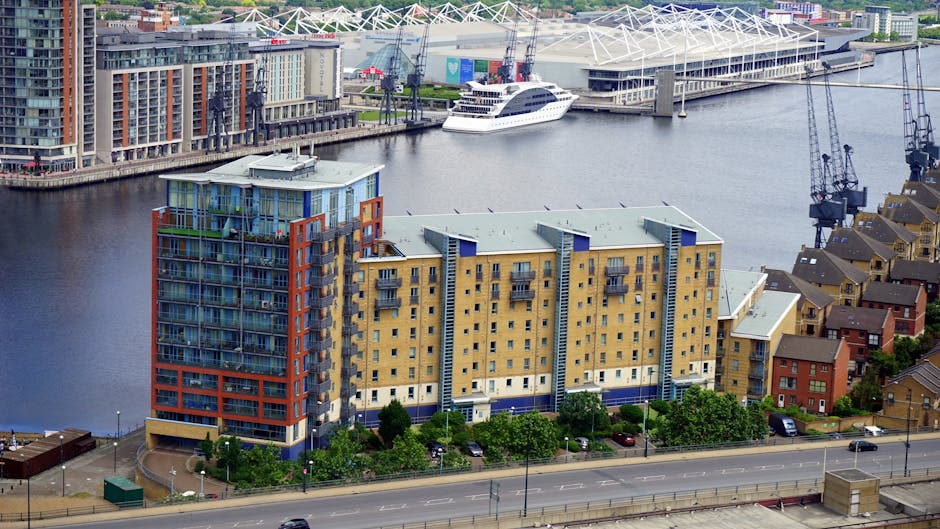 A cityscape featuring modern high-rise buildings along a waterfront, with reflected images of the skyscrapers on the calm water surface. The buildings exhibit a variety of architectural styles, with glass facades, concrete finishes, and some with textured or patterned exteriors. The sky above is clear and blue, and the natural daylight highlights the details of each structure. In the foreground, the water is smooth, creating a mirror-like reflection of the urban skyline. The scene appears to be taken from a quay or promenade area, with no visible waste or clutter, emphasizing the clean, developed environment. The modern appearance of the buildings, combined with the water reflection, suggests a contemporary urban area that could benefit from private waste management or rubbish removal services provided by companies like Rubbish Collection Docklands, supporting effective on-site clearance of waste generated in such metropolitan settings.